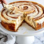 Whole cinnamon roll honeybun cheesecake with glazed top and a slice being removed